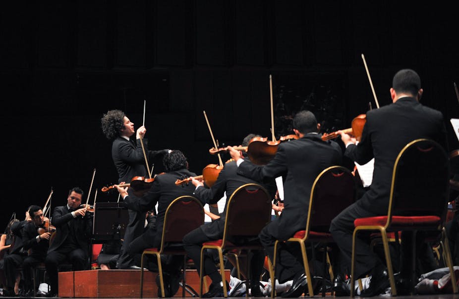 Un hombre con la batuta en alto dirige a muchos músicos sentados empuñando violines.