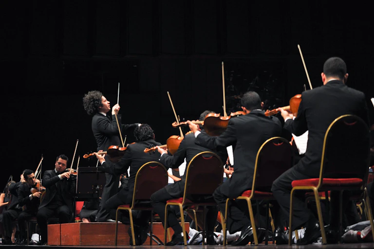 Un hombre con la batuta en alto dirige a muchos músicos sentados empuñando violines.