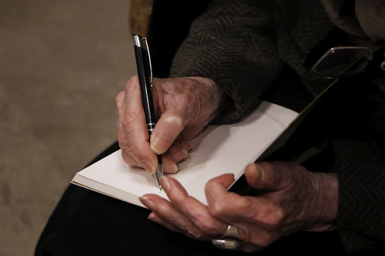 Unas manos ancianas sostienen un bolígrafo antes de escribir en la página en blanco de un libro.