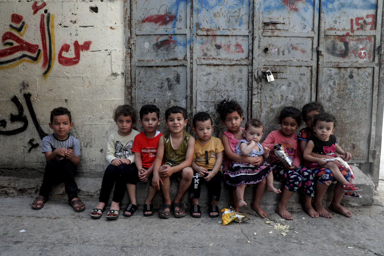 Children sitting in a row on a stone platform next to a large door with a padlock.