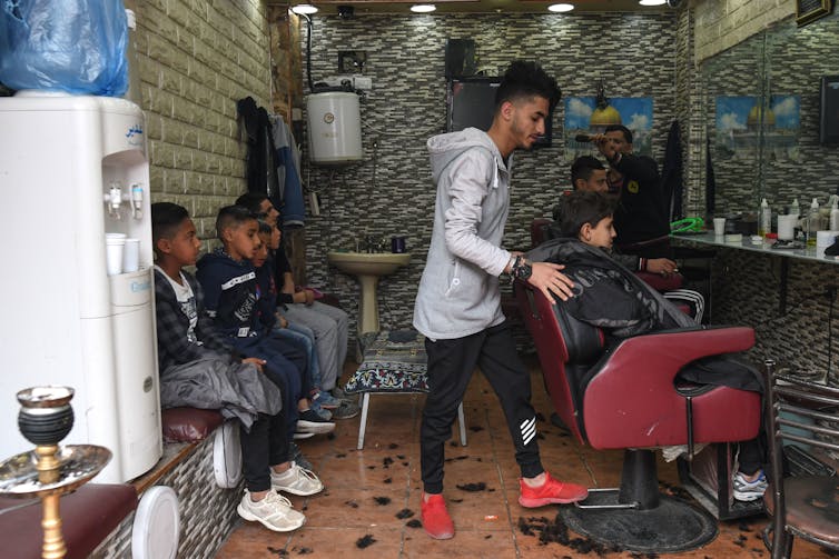 A young barber getting a young child seated on a chair while several others wait.