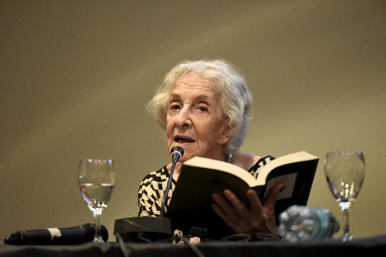 Una mujer delgada y de pelo cano lee de un libro que sostiene en las manos delante de un micrófono.