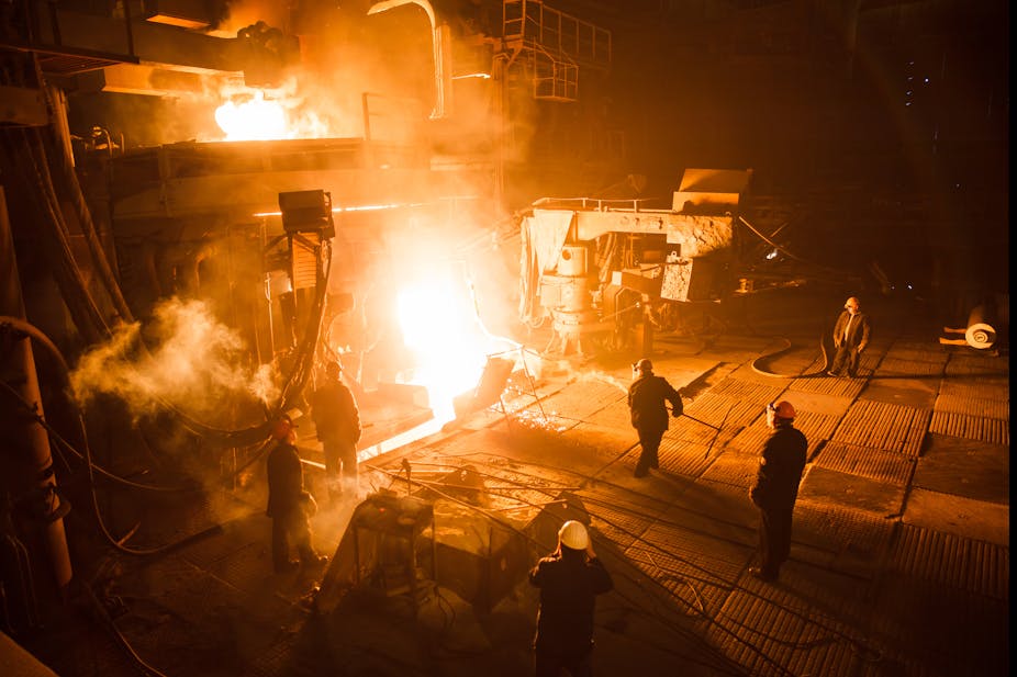 Workers in a steel plant stand around a furnace containing hot metal. 