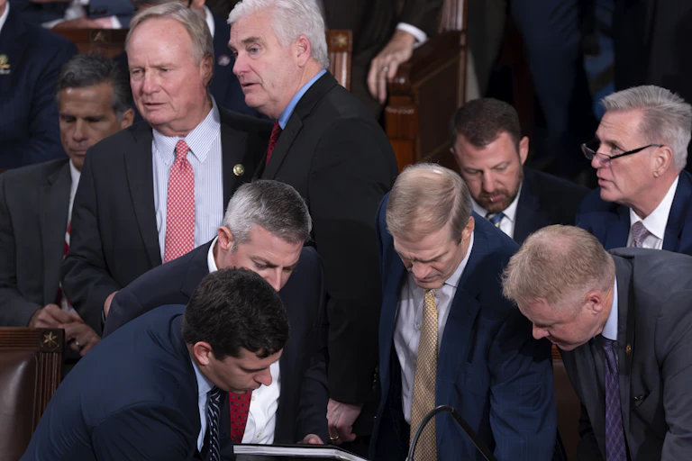 Nine men in suit jackets or blazers, crowded together and looking concerned.