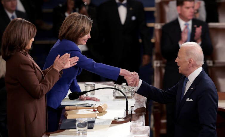 A dark-haired women in a purple jacket shakes the hand of a white-haired man in a suit.