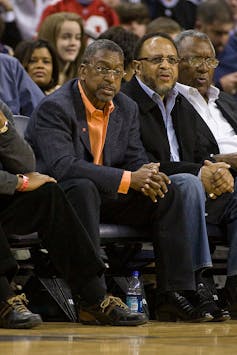 Black man wearing suit jacket and orange shirt seated courtside during a basketball game.