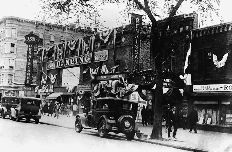 Old photo of street featuring arena with vintage cars parked along the sidewalk.