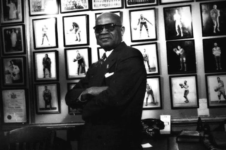 Black bald man wearing sunglasses and a suit poses while folding his arms.