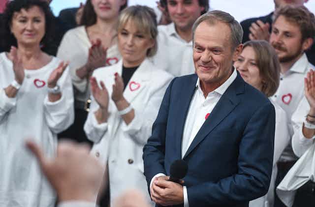 A man in a suit and open-neck shirt stand in front of women dressed in white blazers.