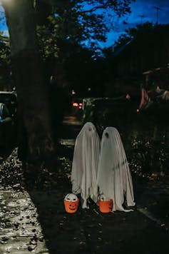 Two kids dressed as ghosts with pumpkin buckets, trick or treating on a street.