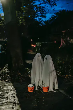 Two kids dressed as ghosts with pumpkin buckets, trick or treating on a street.