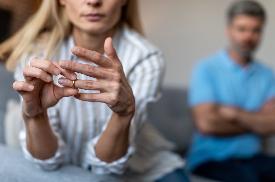 Woman takes off wedding ring sitting on sofa, man with folded arms sits behind her