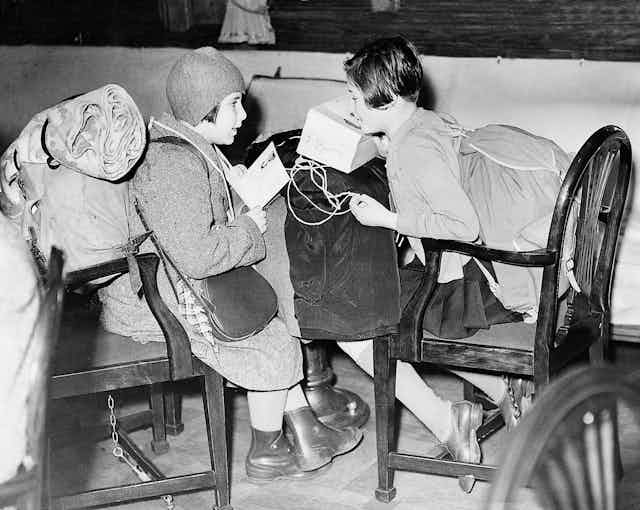 A black and white photo of two children sitting on wooden chairs carrying large bags and sacks. 