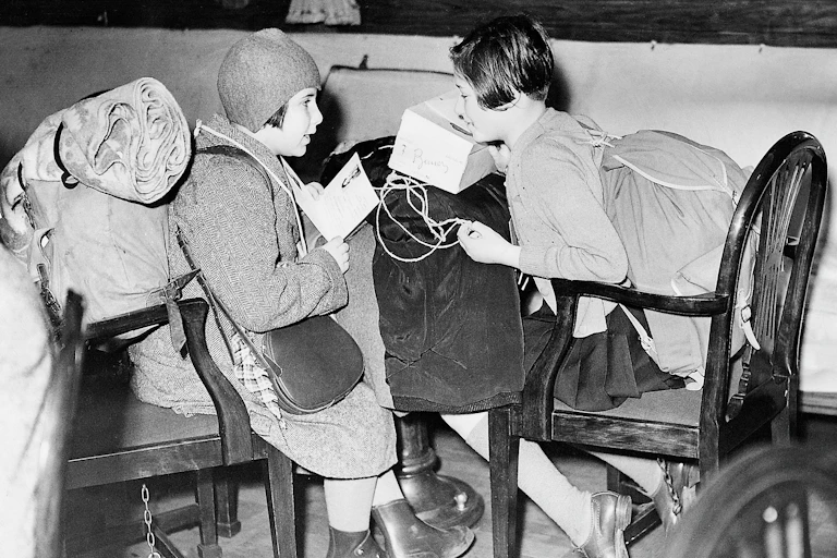 A black and white photo of two children sitting on wooden chairs carrying large bags and sacks.
