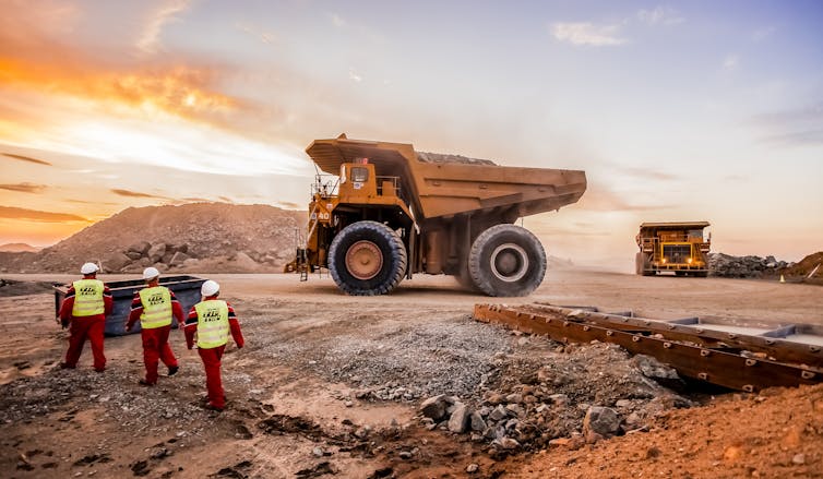 a truck on a pile of rocks and dirt, three people in safety gear are in the foreground