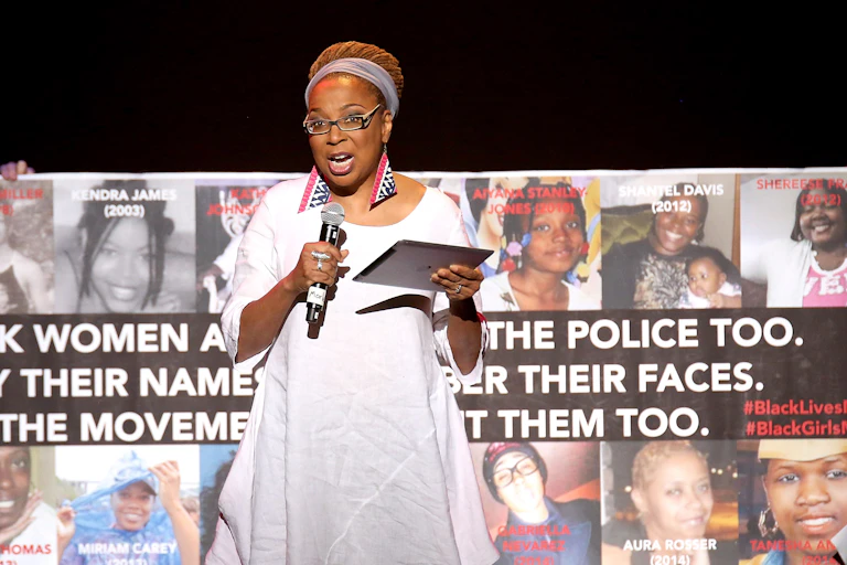A Black woman dressed in a white dress stands on a stage with a microphone in her hand.