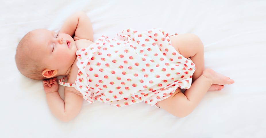 Un bébé habillé d'une barboteuse blanche représentant des fraises dort couché sur le dos, sur un drap blanc.