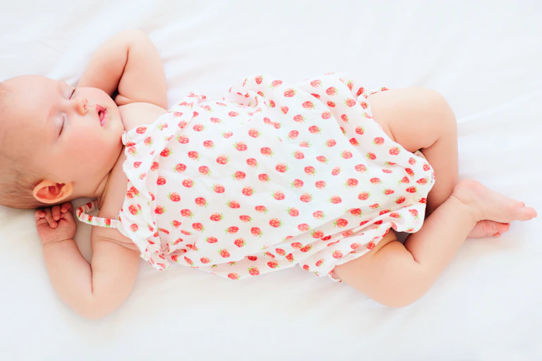 Un bébé habillé d'une barboteuse blanche représentant des fraises dort couché sur le dos, sur un drap blanc.