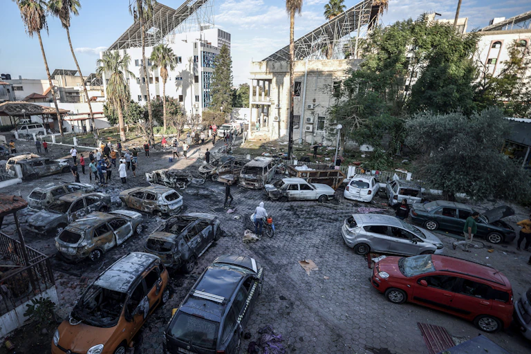Destroyed and burned cars are seen in a parking lot, with palm trees and a white building nearby./