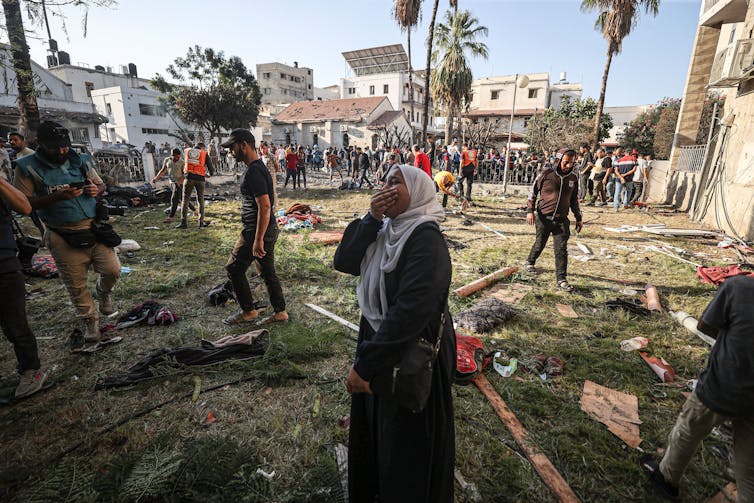 A woman holds her hand over her face and cries in a grassy field that is cluttered with people's belongings. Other people walk nearby.