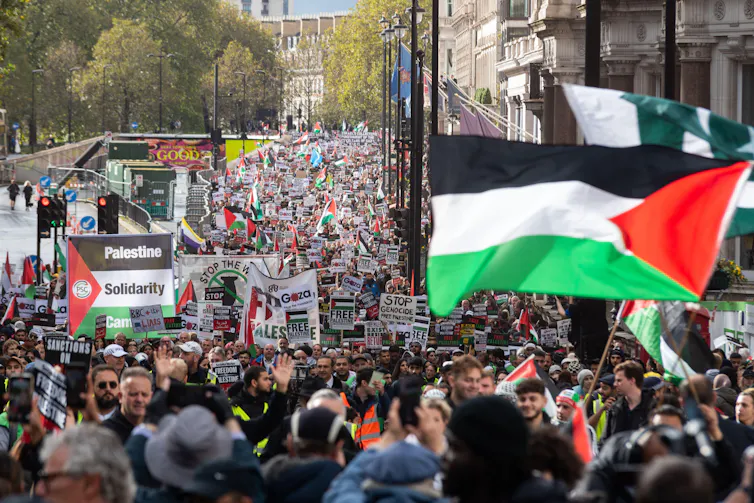 Photo of a large crowd of marchers carrying placards, a large Palestinian flag is in the foreground