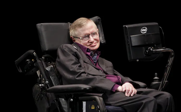 A man wearing a suit sits in an electric wheelchair with a computer screen attached to it.