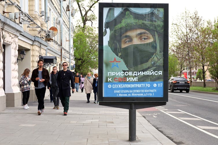 Une affiche dans une rue montre un soldat portant un masque noir et un casque kaki, avec des passants en arrière-plan