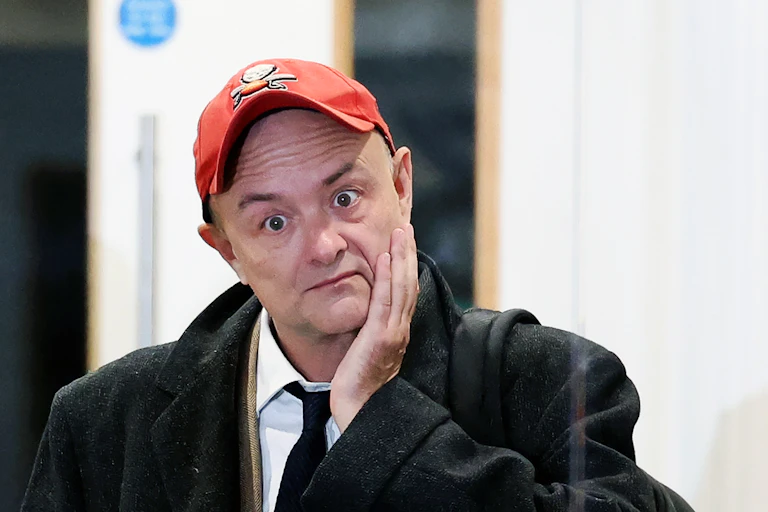 Dominic Cummings wearing a shirt and tie and baseball cap, touch his face and looking wide eyed.