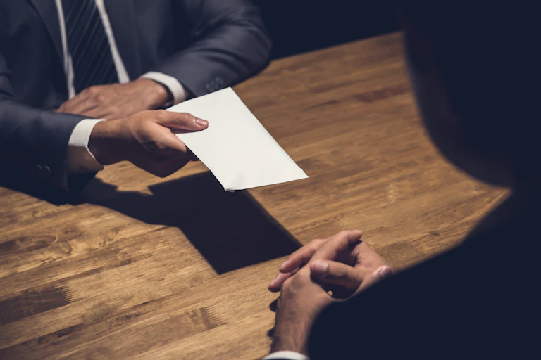 A person, seen from the neck-down, passing a blank envelope to a person sitting with their back to the camera