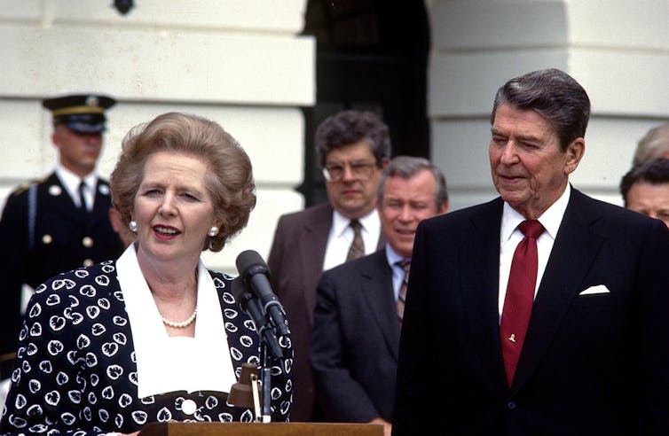 An older woman speaks from behind a podium while an older man in a suit looks on.