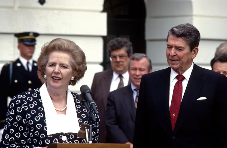 An older woman speaks from behind a podium while an older man in a suit looks on.