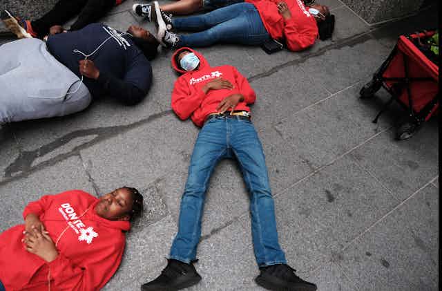 Black teens in jeans, sweats and hoodies lie faceup on a dirty sidewalk with their eyes closed