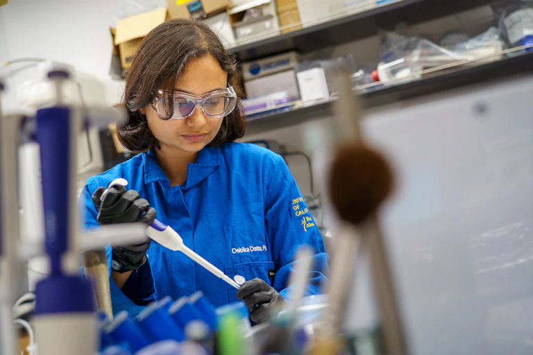 A researcher wearing a blue lab coat and protective goggles holds a white pipette while standing in a laboratory.