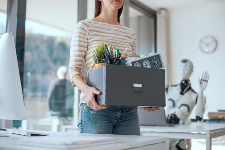 Woman carries a box of her belongings out of an office as a robot waves goodbye to her