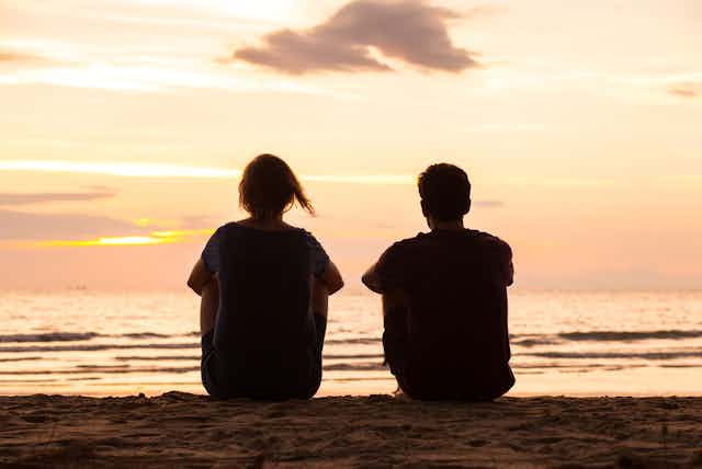 Two young people sitting on beach staring out at sea, sun setting