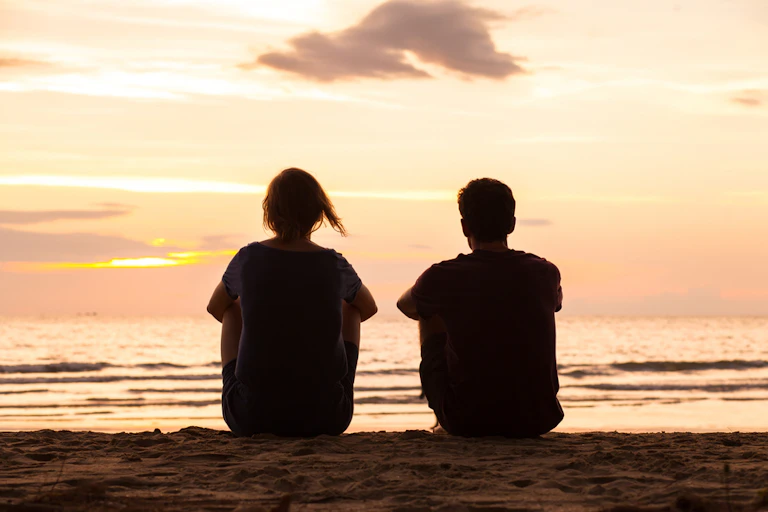 Two young people sitting on beach staring out at sea, sun setting