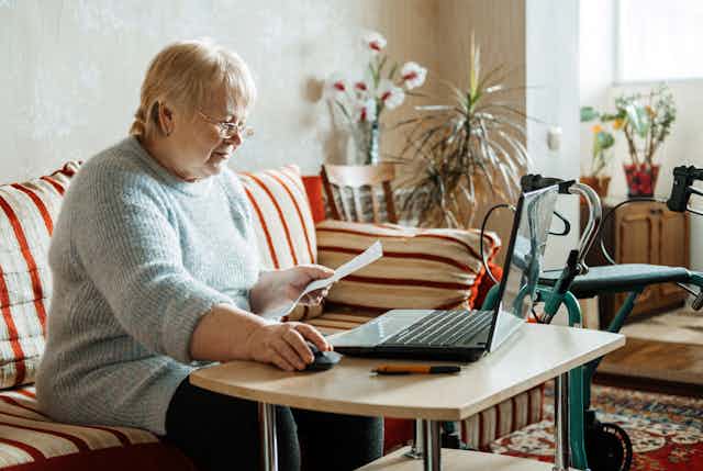 older woman sits at table with laptop, wheeled walking frame nearby