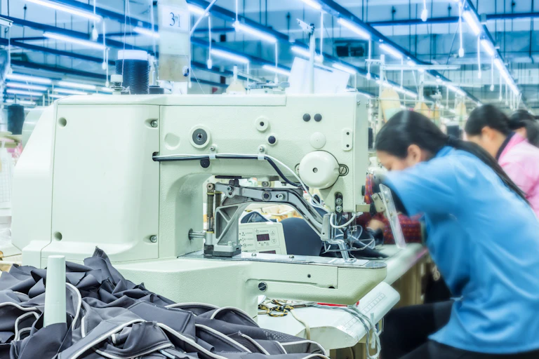 Women sit side-by-side at a row of tables sewing clothing in a factor