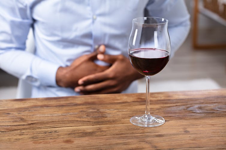 A man clutches his stomach while a glass of red wine sits on a table nearby.