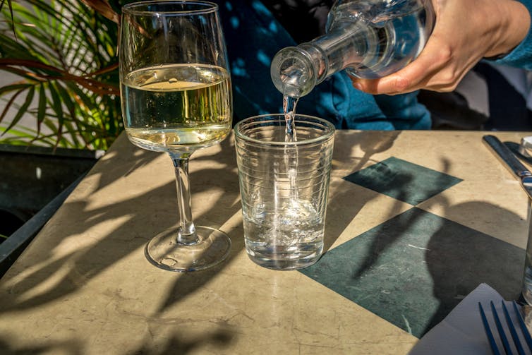 A person pours water into a cup, which is sitting next to a wine glass filled with white wine.