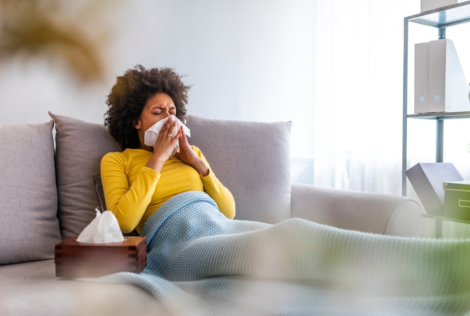 A sick woman reclines on her couch while blowing her nose with a tissue.