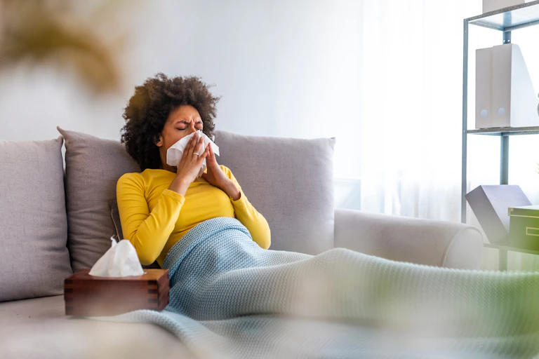 A sick woman reclines on her couch while blowing her nose with a tissue.