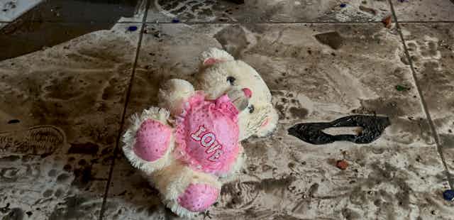 A white teddy bear with a pink heart that reads 'love' lies amid rubble.
