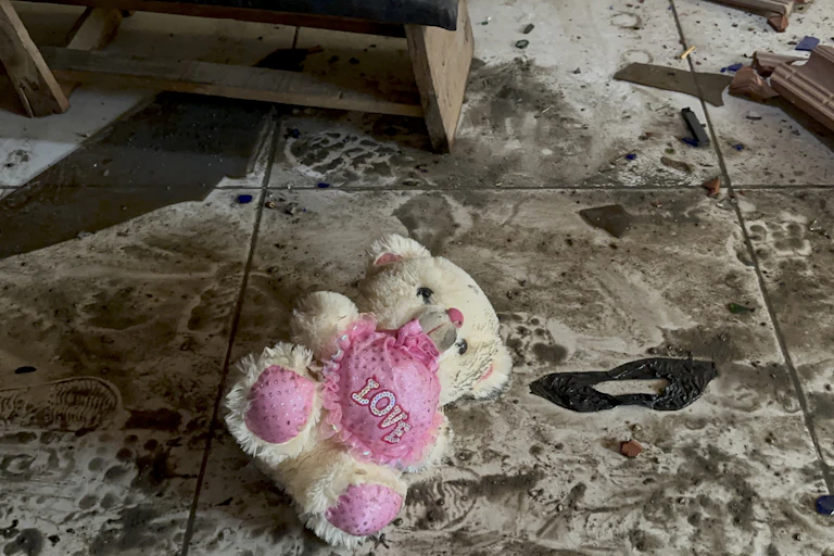 A white teddy bear with a pink heart that reads 'love' lies amid rubble.
