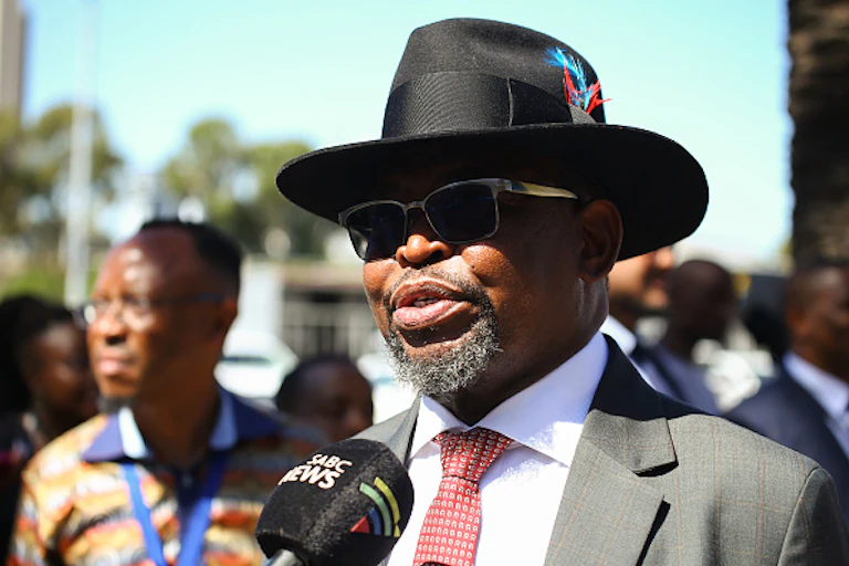 A man in a hat, wearing dark glasses, speaks into a news media microphone.