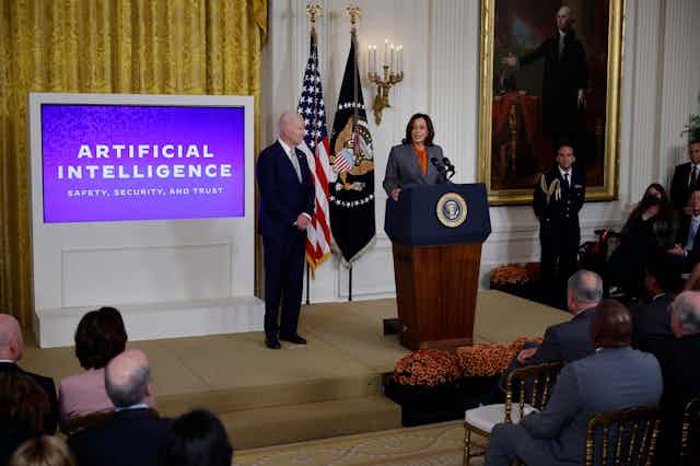 a woman speaks at a podium to a room of seated people while a man stands nearby watching