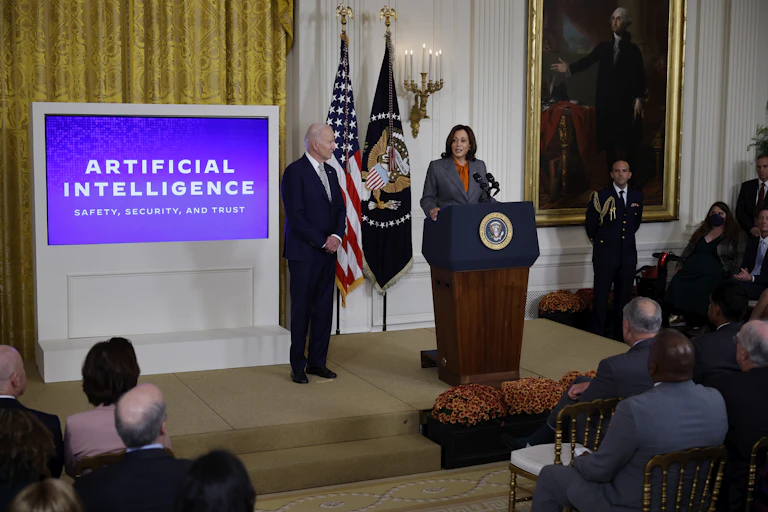 a woman speaks at a podium to a room of seated people while a man stands nearby watching