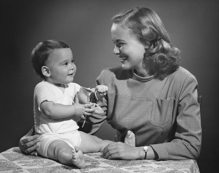 Black and white 1950s photo of a seated baby with a smiling woman