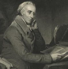 Grey-haired man in jacket sitting at a desk reading,