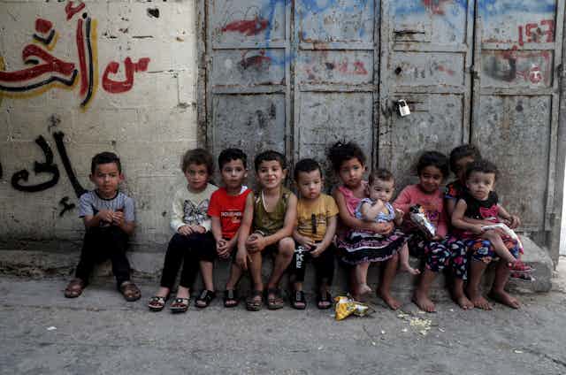 Crianças sentadas em fila em uma plataforma de pedra ao lado de uma grande porta com um cadeado.